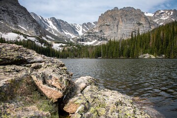 View of Loch Lake in Rocky Mountain National Park in Colorado. A mountain face can be seen as well as a large blue lake and blue skies with clouds and rocks.