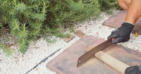 A man sawing cutting wooden joist or plank in a slow motion with hand saw on rock plate at the garden, rusty blade creates a cloud of sawdust while the builder or carpenter is pressing timber to keep	