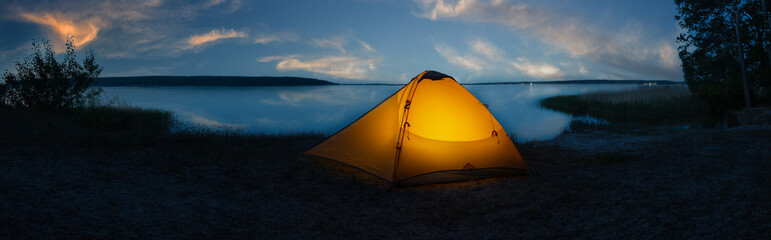 Orange tourist tent illuminated from inside on shore of lake under sunset sky