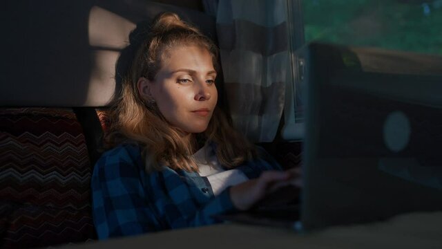 Handheld, female working at a laptop while sitting in a car, typing text and remote work in nature, portrait in a sunset light inside a camping car.