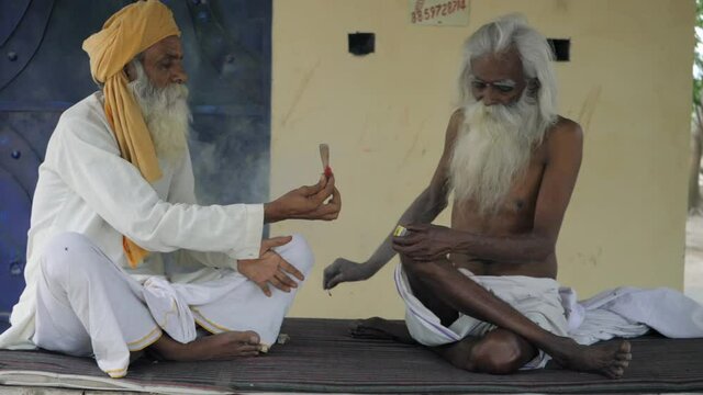 Elderly men in white Kurta Pajama smoking together at home - lifestyle and addiction. Closeup shot of a bare-chested man of a rural Indian village lighting a chillum using a matchstick