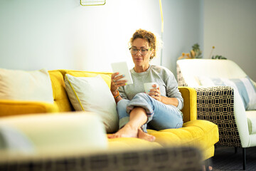 Modern middle age woman read an ebook on tablet sitting comfy on the sofa at home enjoying indoor...