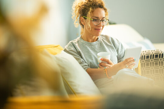 Portrait of beautiful adult woman smile at home sitting on the sofa reading an e-book on online device and enjoy break time - indoor leisure study activity modern female people with glasses