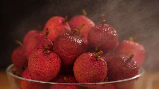 Freshwater is being sprayed / sprinkled on a bunch of red lechees - healthy fruit. Closeup shot of a summer fruit rotating on a wooden table against a dark background