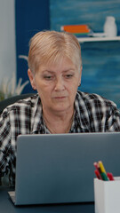 Old middle aged businesswoman working at laptop from home, senior mature woman typing on pc financial data sitting at desk. Employee using modern technology while retired husband sitting on couch