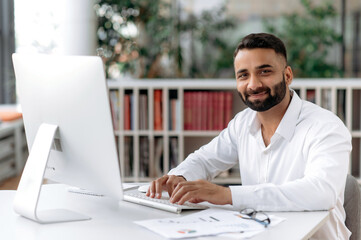 Fototapeta na wymiar Portrait of a handsome confident successful bearded Indian business man, manager, lawyer sitting at work desk wearing white formal shirt, looking straight into camera, smiling pleasantly