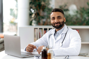 Portrait of an Indian general medicine doctor, sit at a desk in an office in a clinic, dressed in...