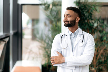 Indian handsome confident general medicine doctor, stands in a clinic, dressed in medical clothes...