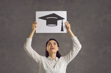 Studio portrait of female student holding paper placard with square academic cap image. Happy young woman looking up at graduate hat picture. Education opportunities, getting university degree concept - obrazy, fototapety, plakaty