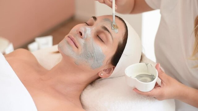 Cleaning and moisturise nourish skin of face. Cosmetologist put grey mask on face by brush. Beautiful woman receiving cavitation facial peeling. Treatment skin care in cosmetology beauty spa salon.