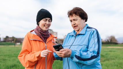 Portrait of smiling adult granddaughter teaches her grandmother how to use a smartphone. Outdoor....