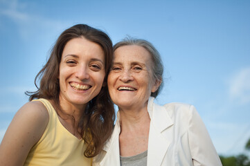 Senior mother with gray hair with her adult daughter looking at the camera in the garden and hugging each other during sunny day outdoors, mothers day