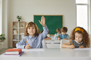 Boy and a girl are sitting at a school desk, writing, the boy raises his hand up. Primary school classmates are sitting in the classroom, the books are on the table, back to school.
