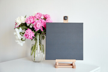Black canvas mockup on wooden easel and vase with pink flowers on table on white wall background