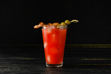 Glass of bloody mary cocktail garnished with bacon and olives on dark wooden background