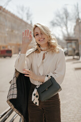 Cheerful young blonde woman in pearl necklace, white blouse and brown pants smiles sincerely, waves...