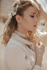 Curly blonde young woman with pearl jewelry and dressed in white blouse looks calm and poses in...