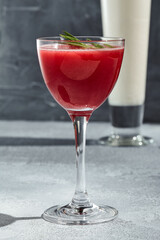 Red Square Cocktail is a classic cocktail with raspberry puree and vodka. Cocktail glass on vintage table with harsh shadow from sunlight. Red cocktail garnished with rosemary sprig.