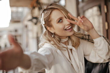 Cool beautiful blonde woman in white stylish blouse and pearl jewelry smiles, shows peace sign and...