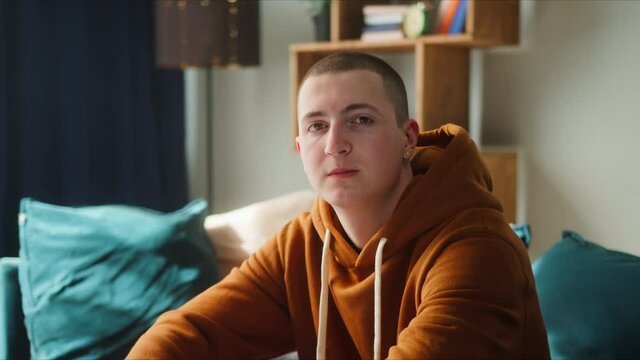 Portrait of brutal teenager with earrings, wearing brown hoodie, young brown-eyed man sitting on sofa and looking in camera with serious face, British model posing. 