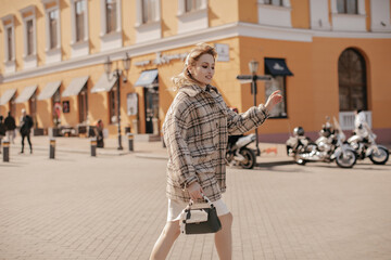 Cool young woman in oversized checkered tweed jacket and white skirt holds black handbag and walks...