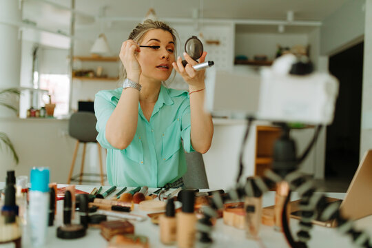 Portrait of young blonde woman blogger recording her new vlog while applying mascara and holding handy mirror. Young woman is recording the makeup tutorial.