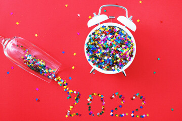 A glass with multicolored sequins, alarm clock and star shaped confettie on the red background. Party, new year, concept