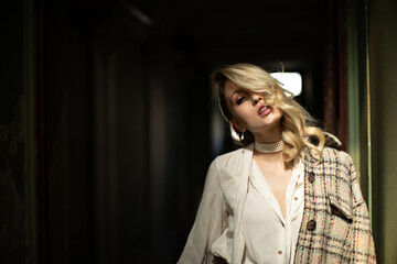 Attractive curly blonde woman in white blouse, checkered jacket and pearl necklace looks into camera and poses in dark room.