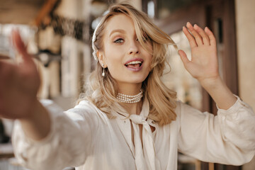 Charming young blonde lady in trendy white blouse and pearl necklace looks into camera, smiles and...