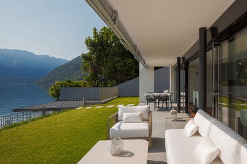 Private terrace overlooking Lake Ceresio in Switzerland. in front of a beautiful green lawn, small table, armchairs and sofa, all white.
