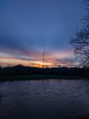 А river and tree branches, then a forest and a radio mast against the backdrop of a sunset and a golden-blue sky.