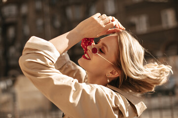 Happy cool blonde woman plays hair outside. Excited lady with red lips in beige trench coat smiles...