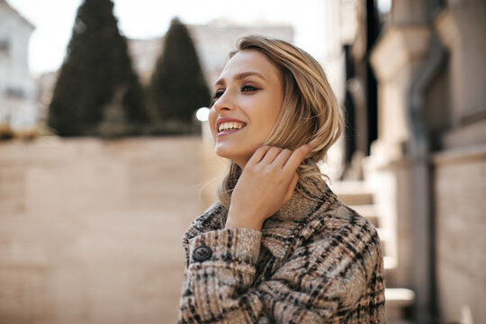 Young beautiful blonde woman in tweed checkered stylish jacket smiles sincerely. Happy lady with beautiful makeup looks away outdoors.