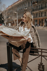 Blonde young curly woman in dark velvet pants, white blouse and checkered coat reads newspaper outside and leans on fence.