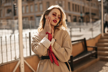 Fototapeta premium Gorgeous blonde elegant woman with red lips in bright colorful sunglasses and beige trench coat gently touches face and poses outside.