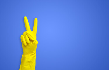 Hand in a yellow rubber glove showing a victory gesture on a blue background with a copy space