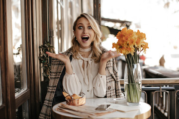 Surprised young woman in white blouse and tweed checkered coat sits in cafe. Attractive girl looks shocked in restaurant.