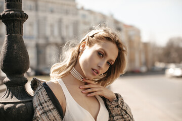 Close-up portrait of blonde gorgeous woman in beautiful makeup looking into camera outside. Attractive lady in white dress and checkered jacket poses at city square.