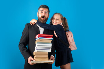 Elementary pupil hugging teacher in studio. Funny father or teacher with school girl daughter hold big stack school textbook notebook books, isolated on blue.
