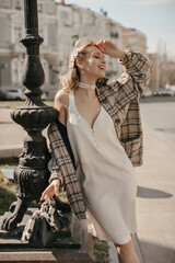 Happy young blonde woman in trendy white dress and tweed checkered coat smiles sincerely and poses...