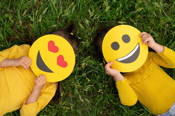 Two children hide behind the faces of cardboard funny happy emoticons lying on the lawn. Top view. World smiley day