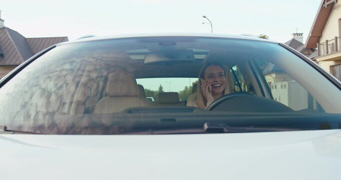 Front view of the happy blonde caucasian woman talking on her smartphone and telling something to her interlocutor while driving a car. People and transport concept