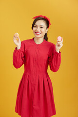 Young asian woman over isolated yellow background holding colorful French macarons and eating it