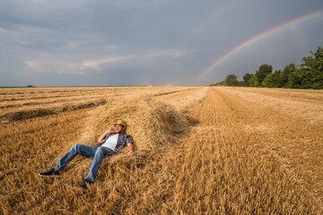 Farmer is resting after successful harvest. Rainbow in the sky behind.