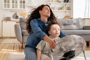 Relaxed mom and little kid sit with closed eyes at fan in living room refreshing from summer heat...