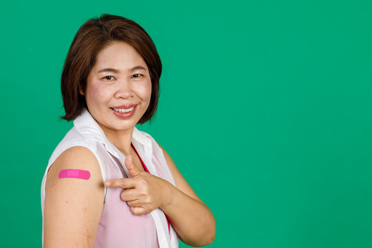 Asian Middle Aged Woman Smiling And Pointing At Her Arm With Bandage Patch Showing She Got Vaccinated For Covid 19 Virus On Green Background. Concept For Covid 19 Vaccination