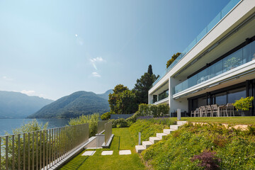 Fototapeta na wymiar Modern house of two apartments with a beautiful garden directly on Lake Ceresio. Sunny day with blue sky. Minimalist and linear architecture. Panoramic view of the lake. Space for your text