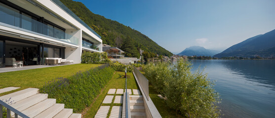 Modern house of two apartments with a beautiful garden directly on Lake Ceresio. Sunny day with blue sky. Minimalist and linear architecture. Panoramic view of the lake. Space for your text