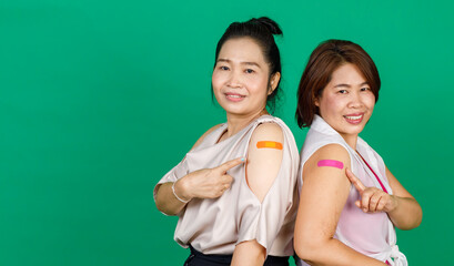 Two middle aged Asian women showing their arms with bandage patch showing they got vaccinated for Covid 19 virus on green background. Concept for Covid 19 vaccination