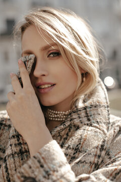 Elegant young blonde woman with beautiful makeup smiles gently, looks into camera and covers part of her face with collar of tweed checkered coat.